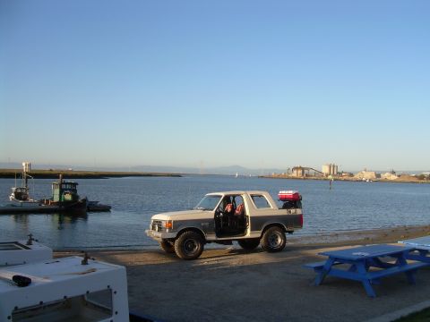 1991 Ford Bronco - A two-tone Ford Bronco is parked near the water with its doors open, carrying luggage on the roof.