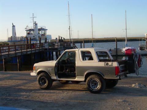 1991 Ford Bronco - A two-tone 1991 Ford Bronco is parked near a dock with boats and a ferry in the background.
