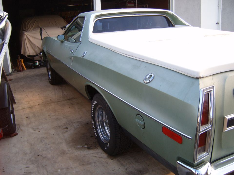 Light green 1973 Ford Ranchero with white bed cover and chrome wheels, rear driver-side view outside a garage