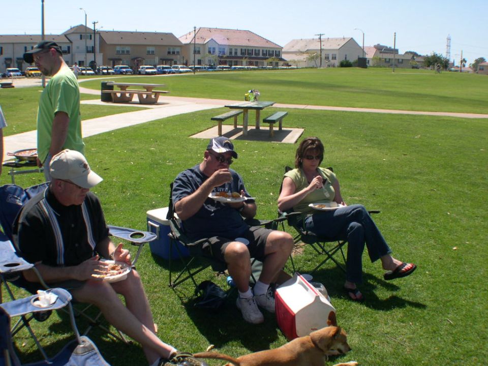 People enjoying a picnic in a grassy park with picnic tables and buildings in the background