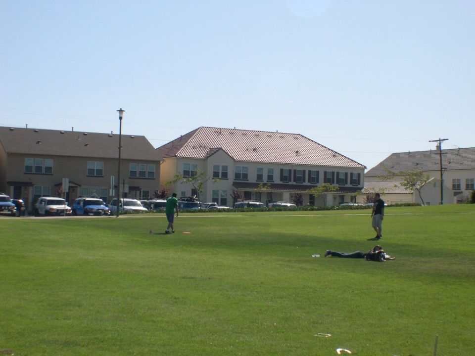 Several cars parked in a lot behind a grassy field with buildings in the background