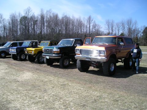 1993 Ford Bronco - A lineup of lifted Ford Broncos are parked on a dirt lot with trees in the background.