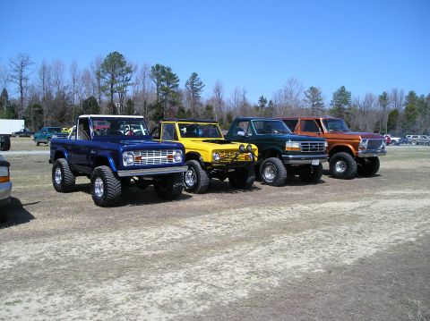 1993 Ford Bronco - A lineup of four modified off-road vehicles, including Broncos and Ford F-150s, are parked on a dirt lot under a clear blue sky.