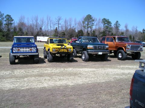 1993 Ford Bronco - Four lifted trucks and SUVs are parked in a line on a dirt lot under a clear blue sky.