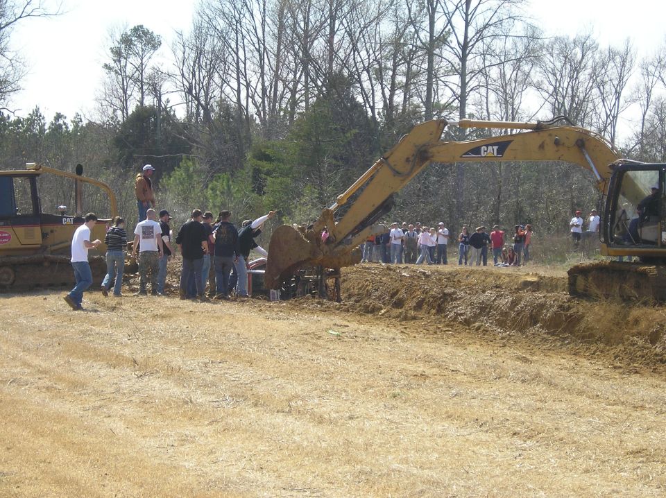 Excavator lifting a car from a ditch with a crowd watching