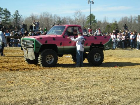 1993 Ford Bronco - A heavily modified pickup truck, painted to resemble a watermelon, sits in a dirt arena with spectators in-ground tires and a crowd in the…