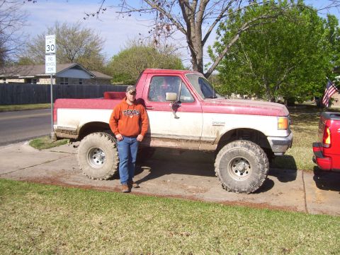 1988 Ford Bronco - A man stands beside a lifted red and white pickup truck, possibly a 1987 model, with large off-road tires.