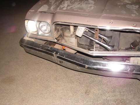 1973 Ford Ranchero - Nighttime front-end view of a tan primer 1970s Ford with the grille removed and the chrome front bumper hanging loose, exposing the radiato…