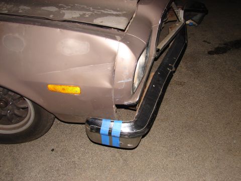 1973 Ford Ranchero - Night close-up of the front corner of a tan/mauve car with the chrome bumper loosened and hanging down, held on with strips of blue painter…