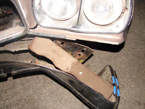 1973 Ford Ranchero - Removed front-end sheet metal laid out on asphalt: a tan header panel section with dual round headlights and chrome bezel, a rusty stamped-…