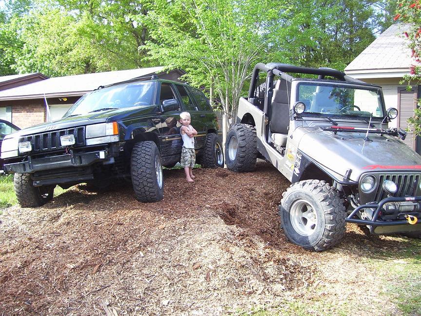 Silver Jeep Wrangler with off-road tires and a black Jeep Grand Cherokee parked on mulch