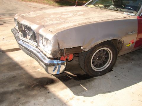 1973 Ford Ranchero - Front-three-quarter view of a faded tan-and-red car-based pickup on a concrete driveway, with the grille and headlight bezel missing, damag…