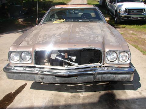1973 Ford Ranchero - Head-on front view of a champagne-tan 1970s Ford with heavily oxidized paint, quad round headlights, and a chrome bumper, its grille remove…