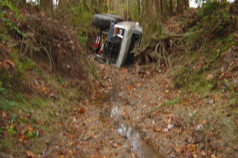 Off-road vehicle flipped onto its side in a wooded ditch with trees and roots around it