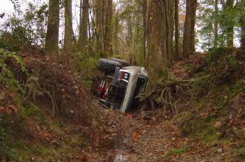 1979 Ford Bronco - A white SUV is overturned in a ditch surrounded by trees and foliage.