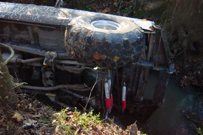 Overturned vehicle with large tire visible, resting in a ditch with water and foliage