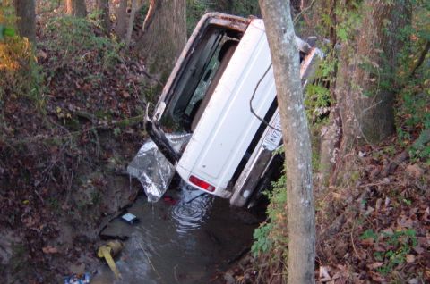 1979 Ford Bronco - A white SUV has gone off-road and is stuck in a water-filled ditch surrounded by trees.