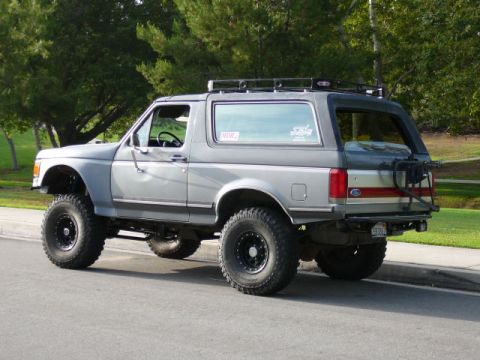 1988 Ford Bronco - This lifted Ford Bronco features off-road tires and a roof rack, parked on a suburban street.