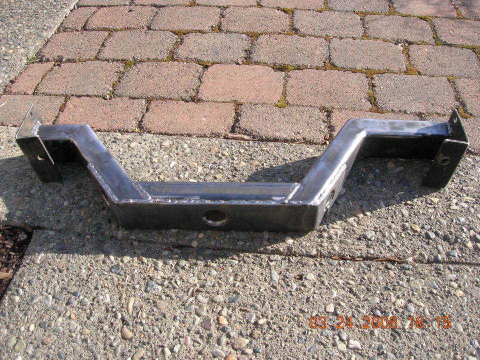 Metal crossmember with welded joints and mounting holes, resting on concrete and brick