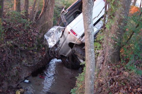 1979 Ford Bronco - A white SUV is lodged in a wooded ditch, its front end heavily damaged.