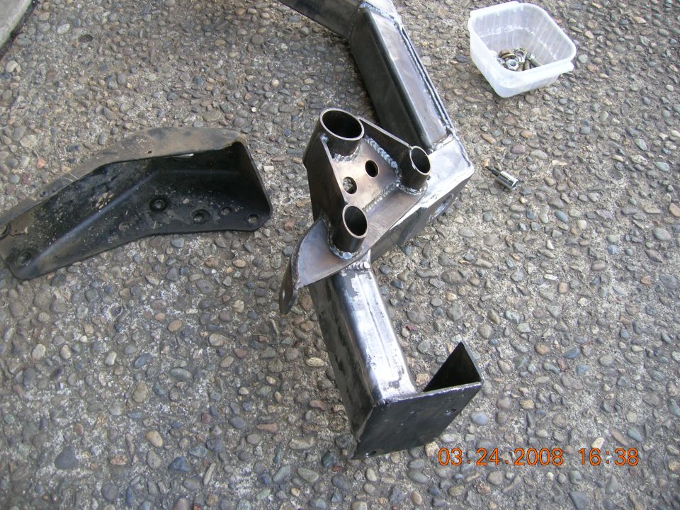 Custom trailer hitch receiver fabricated from steel tubing and plates, with a separate black metal bracket on the left. A small plastic container of nuts and bolts is in the upper right