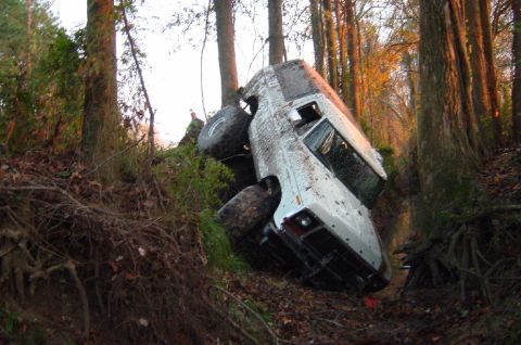 1979 Ford Bronco - White SUV is stuck between trees on a steep, wooded incline.