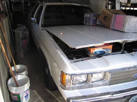 1991 Ford LTD Crown Victoria - A white 1991 Ford Crown Victoria LTD sedan is parked with its hood open, revealing the engine bay and some boxes on the hood.