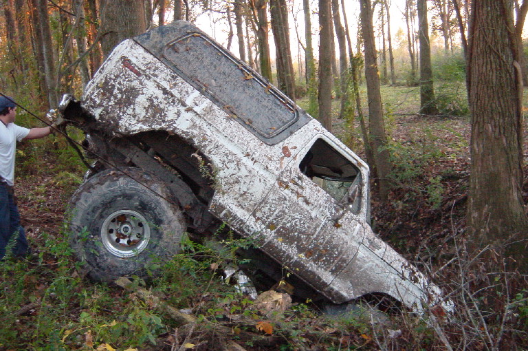 Mud-covered white Bronco tilted nose-down in woods while a person pulls a recovery strap.
