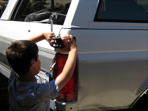 1996 Ford Bronco - A young boy in a blue plaid shirt works on the rear taillight housing of a white Ford Bronco SUV, holding a black light assembly near the e…