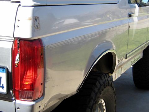 1996 Ford Bronco - Rear driver-side quarter of a silver Ford Bronco SUV, showing the vertical red taillight, a body pinstripe, and dents/scratches along the l…