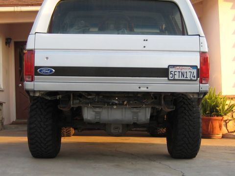 1996 Ford Bronco - Rear view of a white Ford Bronco SUV with a lift kit and large black off-road mud tires, showing the tailgate with a blue Ford emblem and a…
