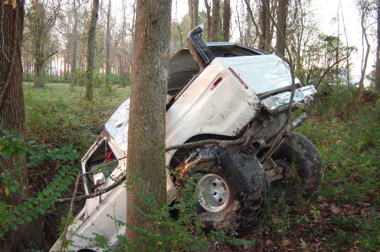 White Ford truck rolled on its side against a tree in the woods, roof crushed, muddy tires up