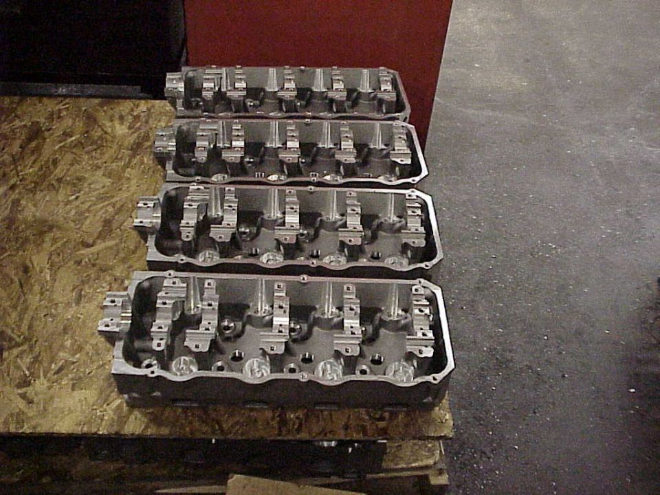 Four bare cylinder heads lined up on a plywood bench with machined aluminum rocker stands installed