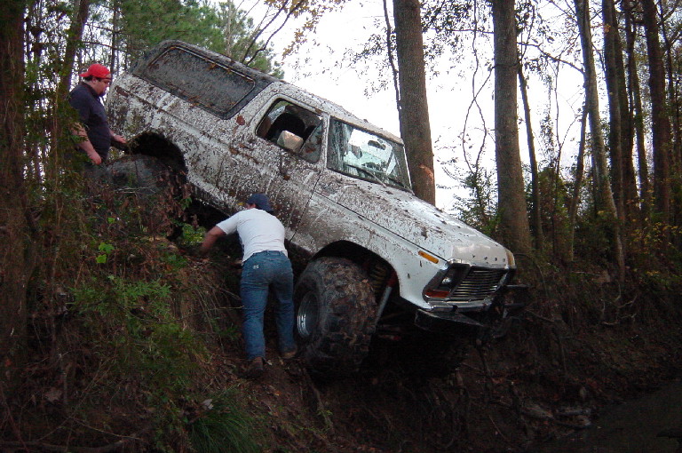Muddy white lifted Ford Bronco tilted on a wooded embankment with two men assisting