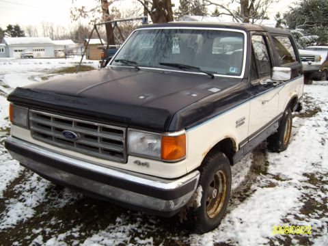 1988 Ford Bronco - A two-tone Ford Bronco sits in a snowy yard, showing its front end and driver's side.