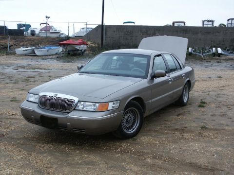 2001 Mercury Grand Marquis - A gray Mercury Grand Marquis is parked in an outdoor lot with boats in the background and its hood up.