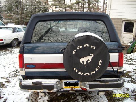1988 Ford Bronco - Rear view of a 1988 Ford Bronco in a snowy setting, showing the spare tire cover and a white car in the background.