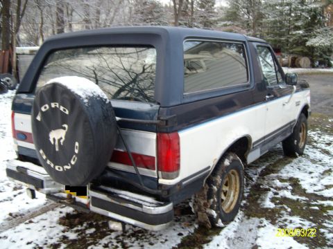 1988 Ford Bronco - Right-rear three-quarter view of a two-tone dark blue over white full-size Ford Bronco SUV parked on a snow-dusted gravel drive, with a bla…
