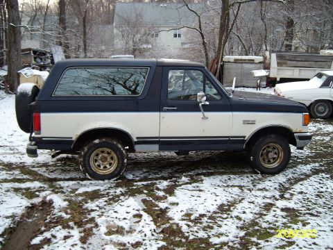 1988 Ford Bronco - Passenger-side profile of a two-tone dark blue over white full-size Ford Bronco parked on snowy ground, showing the removable hardtop with…