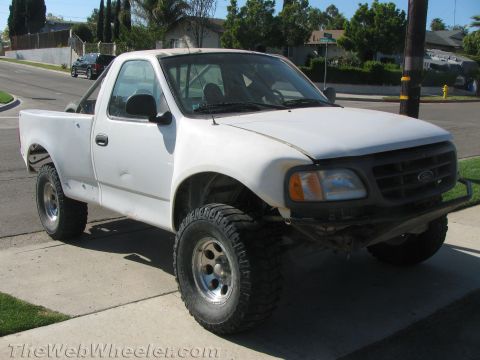 1990 Ford Bronco - white lifted Ford pickup truck with primer-gray fender flares, a tube front bumper, and oversized mud tires parked at a curb.