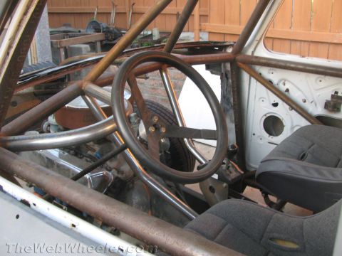 1990 Ford Bronco - Stripped truck cab interior with a bare steering wheel, raw steel roll cage tubing, and a cloth bucket seat during a build.