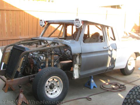 1990 Ford Bronco - partly stripped classic pickup with exposed engine bay, welded roll cage, and Hella lights, sitting on jack stands during a build.