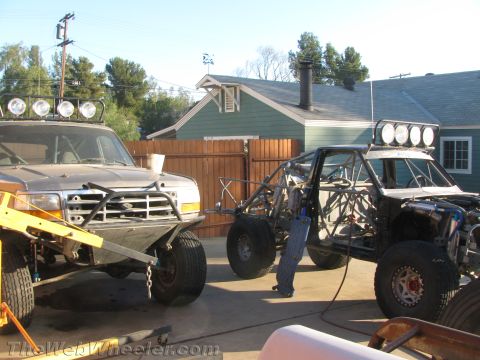 1990 Ford Bronco - lifted Ford truck with light bar parked beside a partly built tube-chassis off-road race vehicle in a backyard work area.