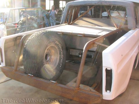 1990 Ford Bronco - Rear view of a stripped pickup race build with a BFGoodrich spare tire mounted in a caged bracket and primer-white body panels.