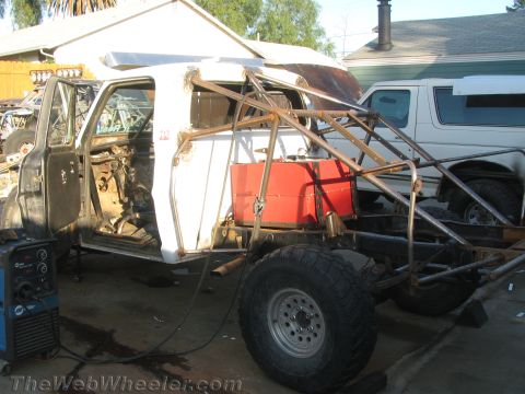 1990 Ford Bronco - stripped pickup cab with welded roll cage, red fuel cell, and bare chassis tubing, with a MIG welder set up in the foreground.