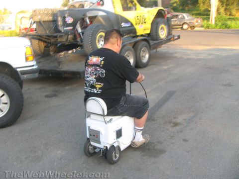 1990 Ford Bronco - man rides a small motorized cooler-scooter in a parking lot behind a truck and trailer loaded with a yellow off-road race truck.