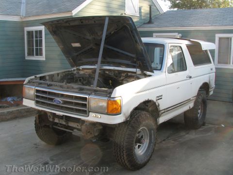 1990 Ford Bronco - white Ford Bronco with its hood raised, lift kit, and oversized tires parked on a driveway next to a teal-green house.