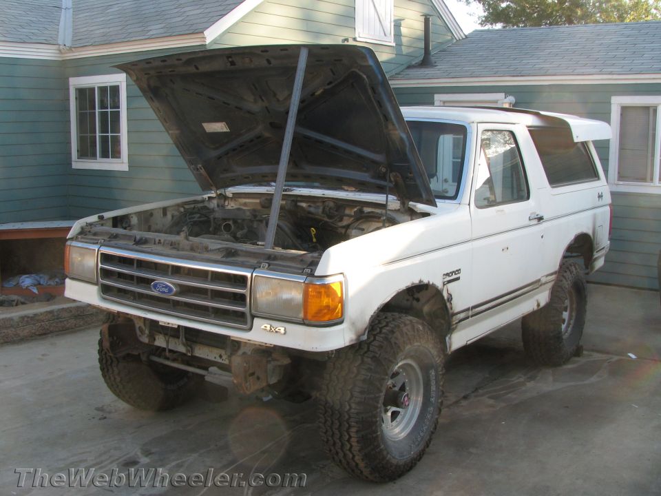 White Ford Bronco with raised hood and lift kit parked beside a green house