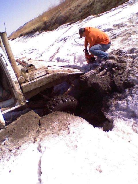 1992 Ford F-350 - A person uses a chainsaw to clear debris around a white truck stuck in a muddy, snow-covered ditch.