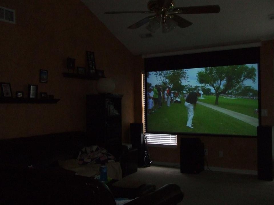 Projector screen shows a golf game with a player swinging. Room has a couch and fan